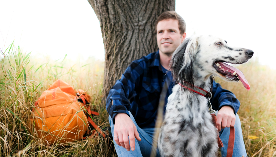 Mand sidder foran et træ med sin orange rygsæk på græsset og hans hund sidder foran ham